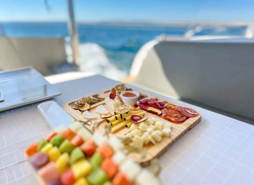 Tábua de petiscos e fruta servida a bordo, com o mar ao fundo